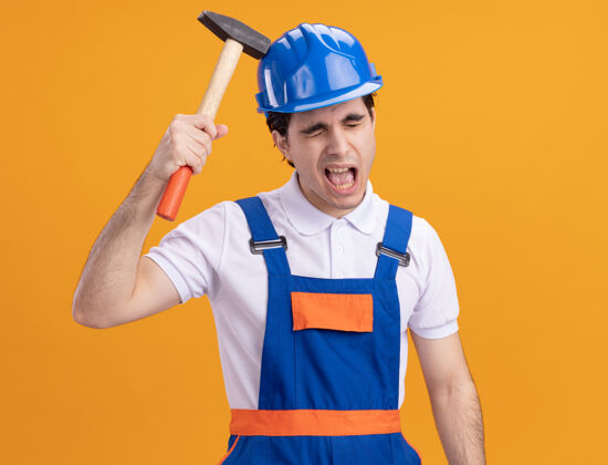 头盔身穿施工制服 头戴安全帽的年轻建筑工人站在橙色的墙上 用锤子敲着自己的头 脸上露出恼怒的表情站着自己男人