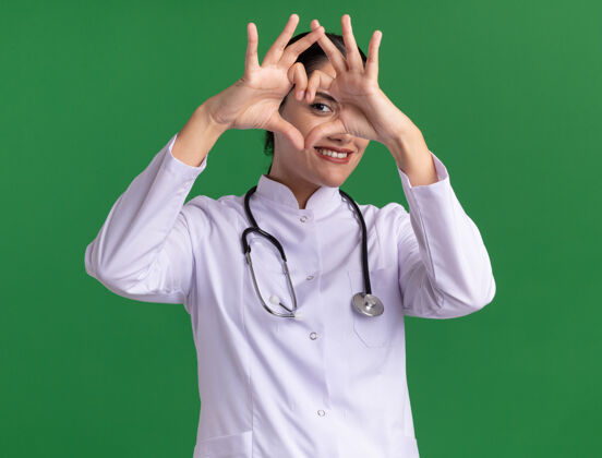 听诊器年轻的女医生 穿着医用外套 手持听诊器 面带微笑 站在绿色的墙上做着手势脸女人站着
