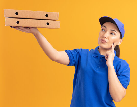 拿着身穿蓝色制服 戴着帽子的年轻送货员站在橙色的墙上 手里拿着比萨饼盒 好奇地看着它们盒子年轻人制服