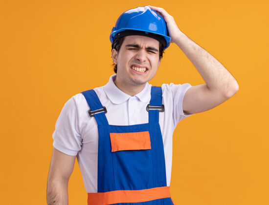 建设者年轻的建筑工人穿着建筑制服 戴着安全帽 看上去很困惑 手放在头上非常焦虑 因为他错误地站在橙色的墙上手头盔制服