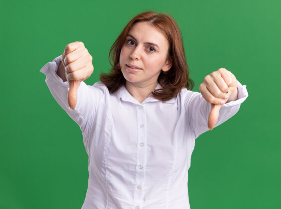 衬衫一位身穿白衬衫的年轻女子 严肃地看着前面 双手竖起大拇指站在绿色的墙上站着女人脸