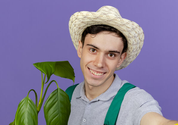 假装微笑的年轻男园丁戴着园艺帽站在植物旁假装拿着相机帽子紫色立场
