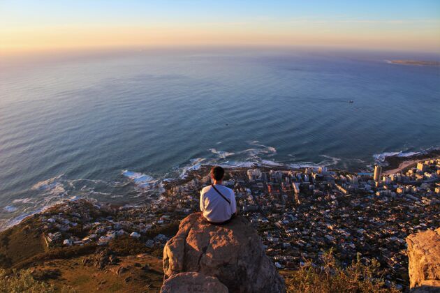 日落横拍一个男人坐在岩石边上 看着城市的海岸线水山天空