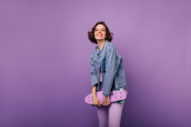 快乐快乐的欧洲女人穿着紫色裤子和滑板合影室内拍摄迷人的微笑女孩 深色波浪发年轻时尚无忧无虑