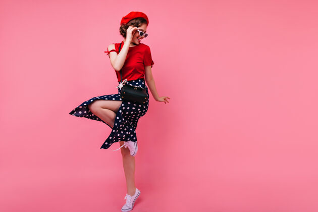 舞蹈兴奋的黑发女士穿着黑色裙子在玫瑰色的墙上跳舞迷人的白色女孩在法国贝雷帽跳跃配件室内时尚