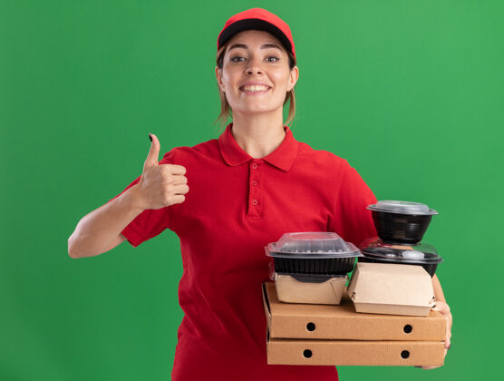 比萨饼微笑着的年轻漂亮的送货女孩 穿着制服 竖起大拇指 拿着纸食品包装和比萨饼盒上的容器拇指容器制服