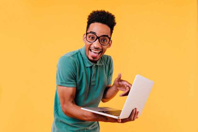 表情一个穿着绿色t恤的学生拿着笔记本电脑摆姿势令人惊讶的男自由职业者的室内照片黄色休闲明亮