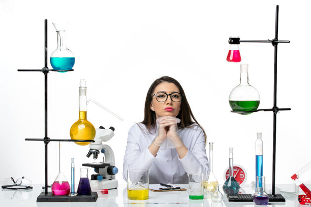 玻璃前视图穿着白色医疗服的女化学家坐在白色背景的科学病毒柯维大流行实验室里 拿着不同的溶液实验室大流行套装