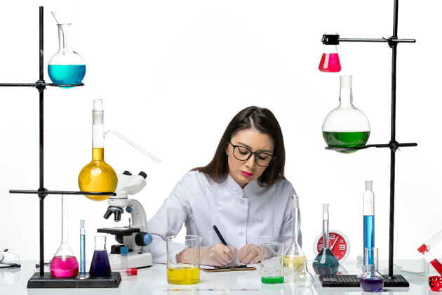 科学前视图穿着白色医疗服的女化学家正坐在那里 拿着解决方案 写着关于白色背景科学大流行实验室病毒covid的笔记实验室视图医学