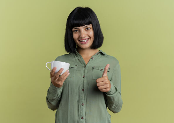 拇指微笑的年轻黑发白种女人拿着杯子 竖起大拇指 在橄榄绿的背景上与复制空间隔离橄榄微笑女人
