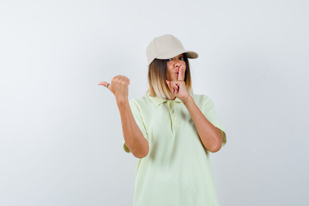 时尚年轻女士展示沉默的手势 同时用拇指指着左边 穿着t恤 戴着帽子 看起来很自信 正面视图成人前面左边