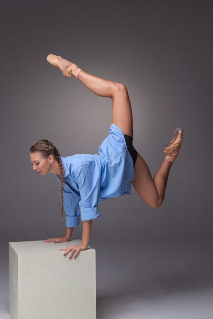 动作年轻漂亮的现代风格舞蹈演员在白色立方体上摆姿势 背景是灰色的工作室女性能量模型