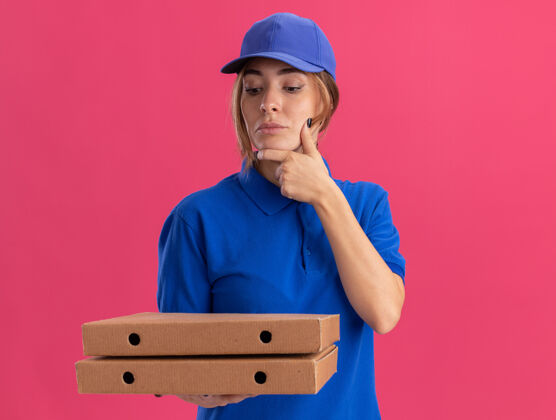 年轻体贴的穿着制服的年轻漂亮送货女孩把手放在下巴上 看着粉红色的比萨饼盒送货思想制服