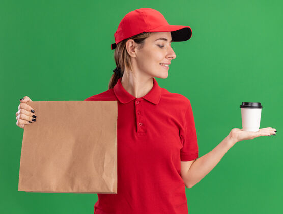 送货穿着制服的年轻漂亮的送货女孩拿着纸包 看着绿色的纸杯姿势年轻成人