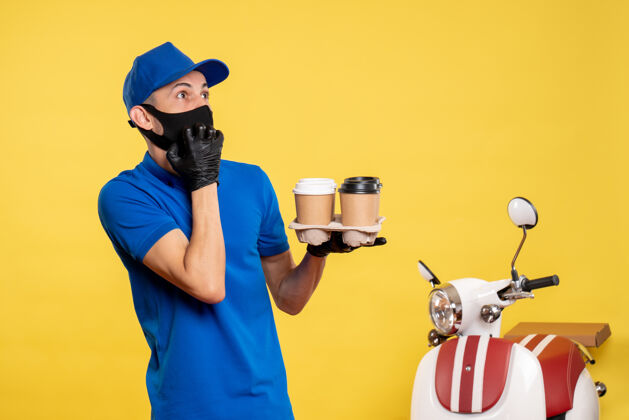 专业正面图：戴着黑色口罩的男快递员拿着咖啡在黄色的工作服上递送工作服持有工作面具