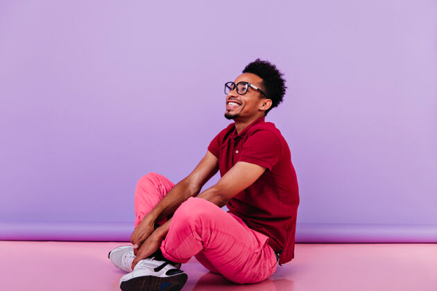 模特笑容可掬的乐观男人穿着粉色裤子坐着情绪激动的黑人年轻人在地板上摆出幸福的微笑时尚有趣美国