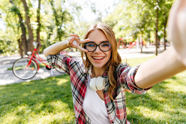 自然戴着眼镜的可爱女孩在公园里自拍夏天 一个漂亮的金发女郎戴着耳机在户外拍摄玩夏天森林