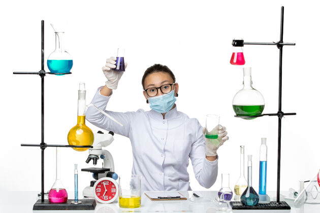持有前视图穿着医疗服的女化学家 戴着面罩 在白色背景上拿着不同的溶液 飞溅着病毒化学实验室的covid-面罩化学研究