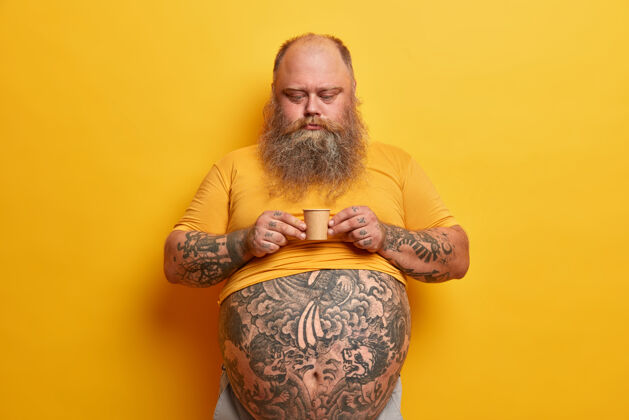 热大肚子 手臂和腹部都有纹身的严肃的大胡子男人 拿着一小盒含糖的咖啡 喝着芳香的饮料 穿着黄色t恤 独自在室内摆姿势男人饮用胡子