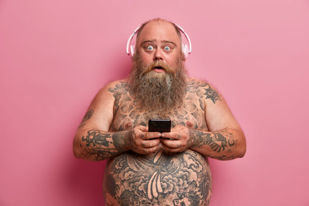 男人惊讶的胖男人盯着他说哇 通过智能手机和朋友发短信 戴着耳机 听音乐 赤膊上阵 纹身科技和休闲理念厚肚子脂肪
