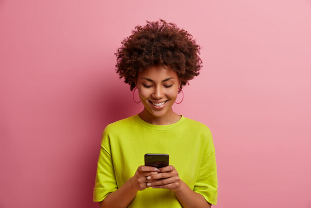 室内高兴的卷发女人在匆忙中输入信息 玩有趣的新游戏 上网发帖 浏览互联网 穿绿色t恤 对着粉色墙壁摆姿势 在移动计划者中做计划罗西社交通话