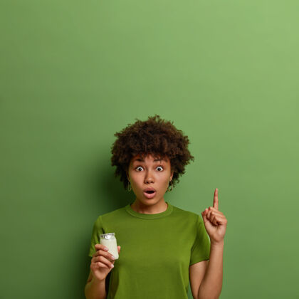 单色情绪激动的卷曲的年轻女子用食指向上指 展示上面令人惊奇的东西 摆出酸奶的姿势 有适当的营养 保持健康的饮食 穿着鲜艳的绿色t恤新鲜早餐卷发