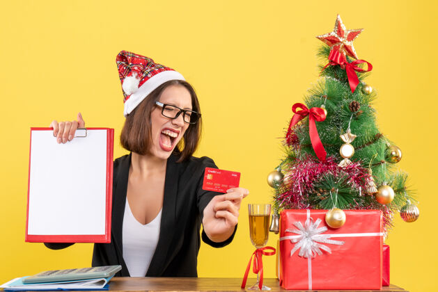 帽子性感迷人的女士穿着西装 戴着圣诞老人的帽子和眼镜 在黄色的办公室里展示着银行卡和文件快乐圣诞老人圣诞老人