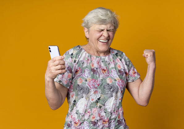 抱着快乐的老妇人举起拳头 手里拿着橘色墙上孤立的电话橘子举起女人