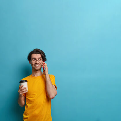 咖啡面带微笑的家伙坐在耳边 忙着和朋友聊天 讨论好消息 端着外卖咖啡 愉快地交流 在蓝色背景下摆姿势 为你的信息复制空间站立帅气情绪