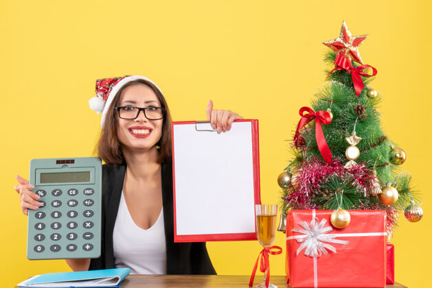 人微笑迷人的女士穿着西装 戴着圣诞老人的帽子 展示着文件 手里拿着计算器 在黄色的办公室里与世隔绝黄色持有人