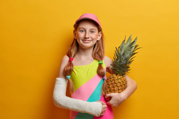 水果可爱的小女孩 穿着五颜六色的泳衣 戴着帽子 拿着菠萝在黄色的墙上摆姿势 享受夏天的时光 好好休息 从高处摔下来摔断了胳膊 或者是危险的运动姿势狐狸成人