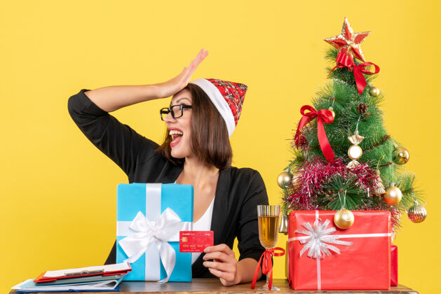 圣诞老人惊喜迷人的女士穿着西装 戴着圣诞老人的帽子和眼镜指着礼物和银行卡在黄色的办公室里孤立人人微笑
