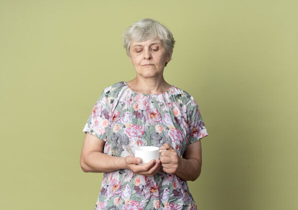 杯子自信的老妇人拿着橄榄绿墙上孤立的杯子看着自信绿色老年人
