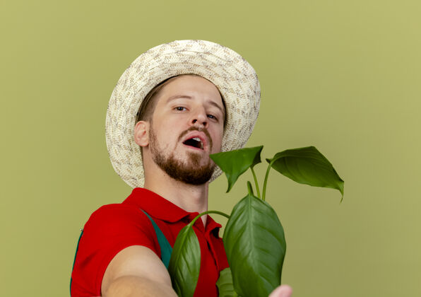 伸展特写镜头中自信的年轻英俊的斯拉夫园丁身穿制服 手持帽子 向孤立的植物伸出手来斯拉夫英俊制服
