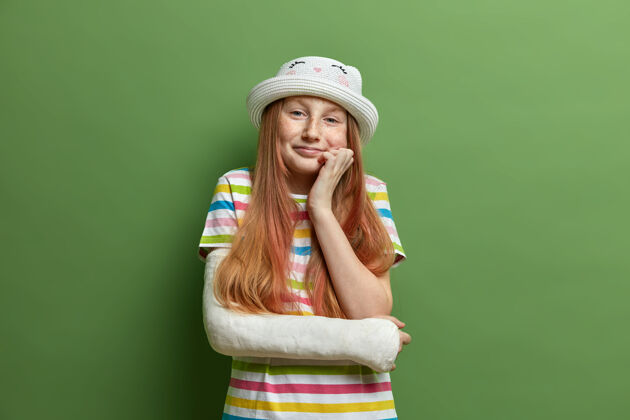 孩子可爱可爱的小女孩把手放在下巴下 有微笑的表情 穿着夏装 意外康复 手臂骨折 看望外科医生后戴石膏意外帽子红发