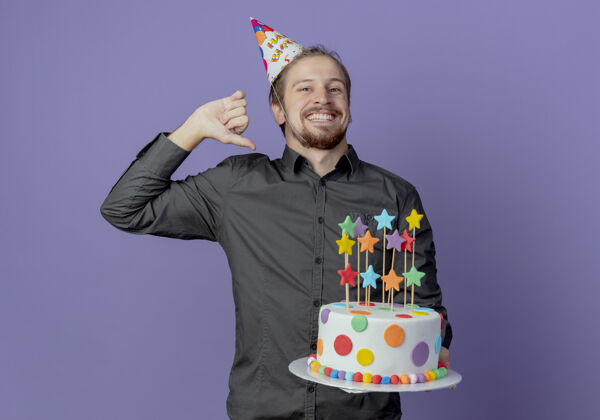 蛋糕戴着生日帽的快乐帅哥拿着并指着隔离在紫色墙上的蛋糕男人生日抱着