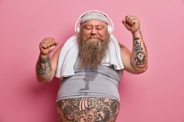 身体快乐的男胖子喜欢音乐锻炼 举手跳舞 全身出汗 脖子上围着衬衫和毛巾 隔离在粉红色的墙上胖乎乎的运动员很高兴取得好成绩大背心减肥