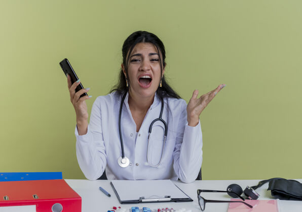 工具愤怒的年轻女医生穿着医用长袍 手持听诊器坐在办公桌旁 手持医疗工具 手持手机 神情显得空手孤立抱着空医疗