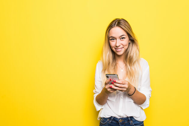 沉思一个沉思的年轻女孩拿着手机 站在黄色的墙上 孤立地看着远方的画像微笑时尚女性