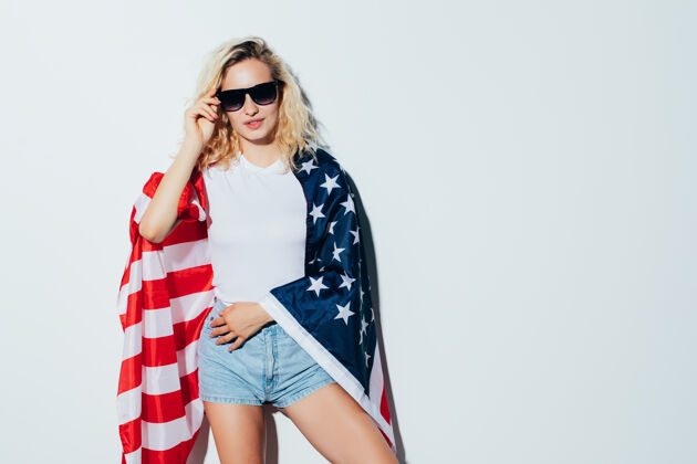 人年轻而无忧无虑年轻漂亮的金发女郎举着美国国旗 微笑着站在白墙上庆祝7月4日独立日模特无忧无虑美国