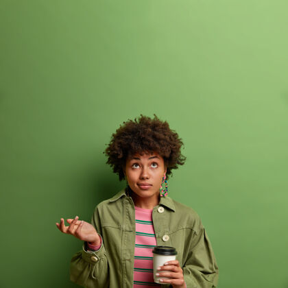 表情迷茫犹豫不决的年轻女子照片在迷茫中抬起手掌 聚精会神地放在上面 喝外卖咖啡 站在室内 穿着时尚的衣服 隔离在绿墙上 复制空间穿思想咖啡