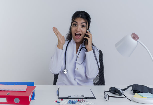 女性令人印象深刻的年轻女医生穿着医用长袍和听诊器坐在办公桌旁 手持医疗工具看着电话交谈 显示空手孤立印象长袍桌子