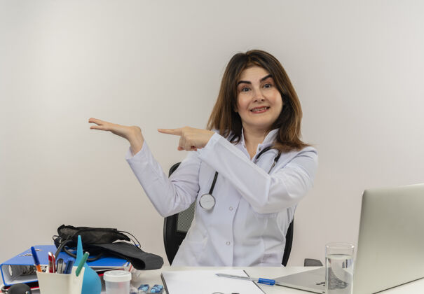 坐着微笑的中年女医生穿着医用长袍和听诊器坐在办公桌旁 手里拿着医疗工具剪贴板和笔记本电脑 手空空的 孤立地指着它笔记本电脑长袍手