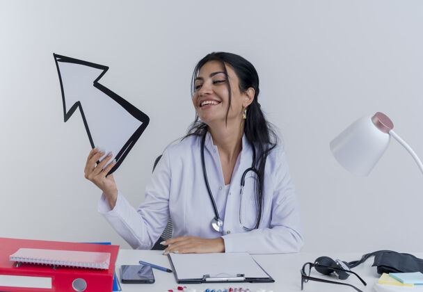 工具快乐的年轻女医生穿着医用长袍和听诊器坐在办公桌旁 手里拿着医疗工具 看着指向一旁的箭头抱着医生女人