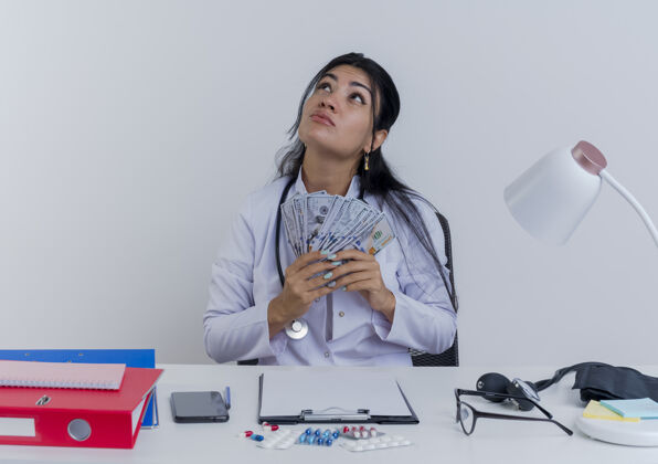 站着体贴的年轻女医生穿着医用长袍和听诊器坐在办公桌旁 手里拿着医疗工具 手里拿着钱 孤零零地抬头看桌子抱着工具
