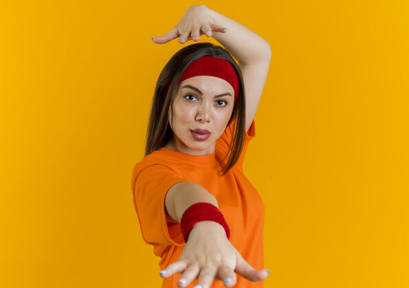 橙色戴着头带和腕带的自信的年轻运动型女人伸出一只手 看着头上的另一只手戴朝向另一个