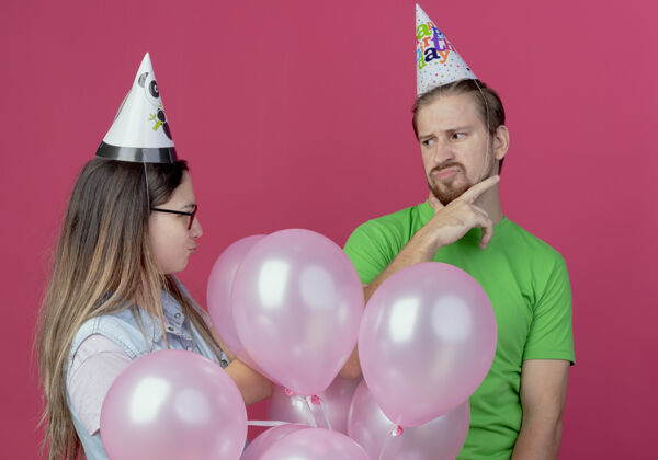 生日戴着派对帽的年轻夫妇不高兴地看着彼此站在粉红色的墙上 氦气球被隔离了年轻站派对