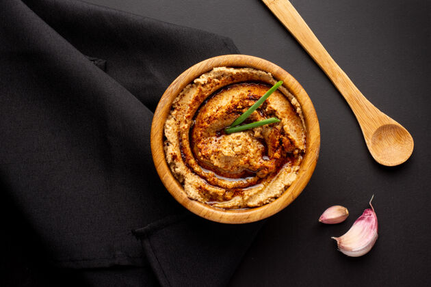 传统头顶拍摄的鹰嘴豆泥在一个木碗里 木勺和大蒜片放在黑桌子上素食配料碗