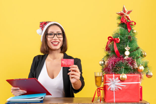 肖像快乐迷人的女士穿着西装 戴着圣诞老人的帽子和眼镜 在办公室里展示着银行卡人西装漂亮