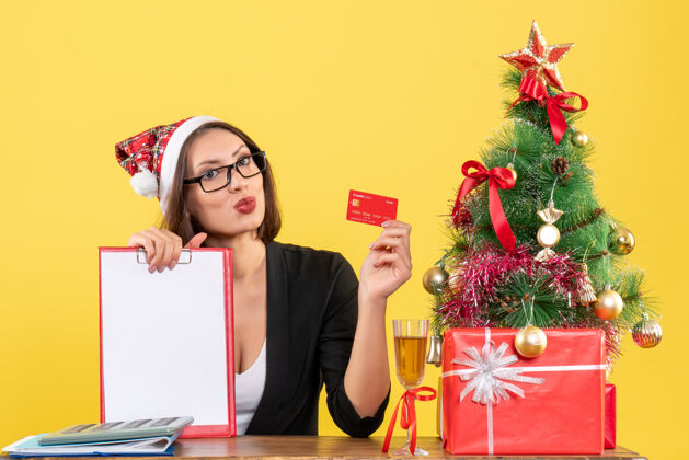 表演性感迷人的女士穿着西装 戴着圣诞老人的帽子和眼镜 在黄色的办公室里展示着银行卡和文件圣诞老人情绪圣诞老人
